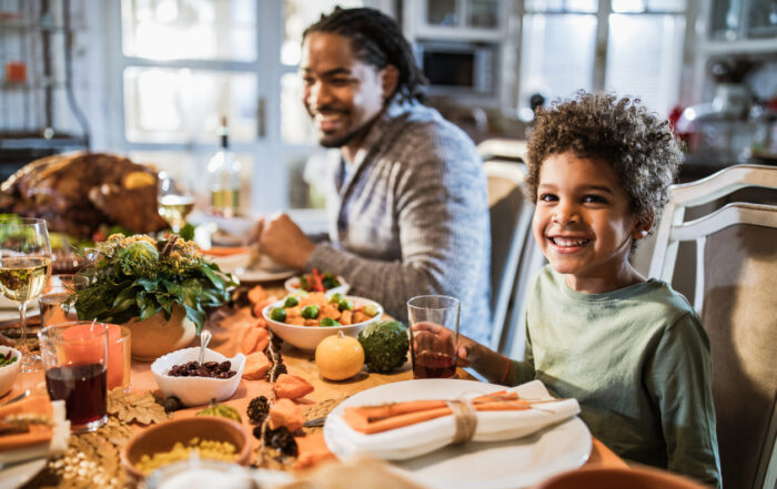 Happy African American boy looking at camera while having Thanksgiving lunch with his father at dining table.